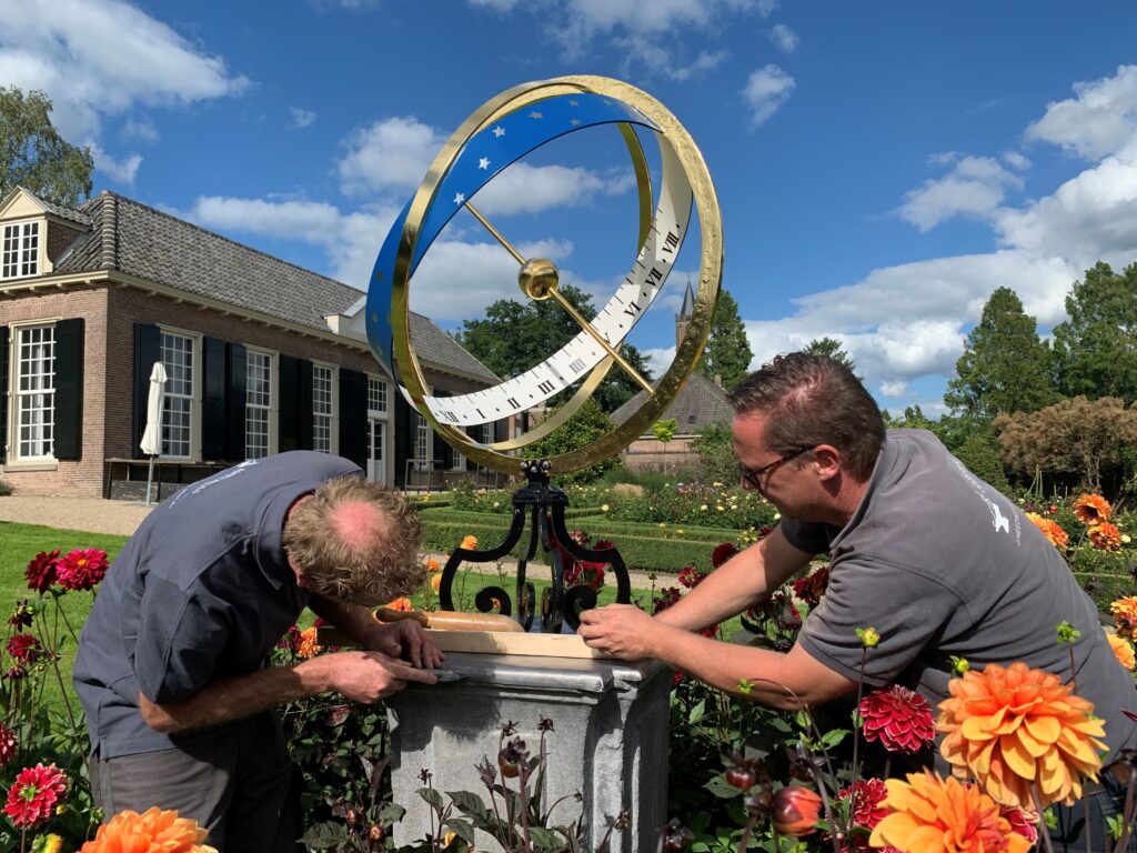 Twee mensen plaatsen de zonnewijzer van Kasteel Amerongen terug op de sokkel in de tuin 